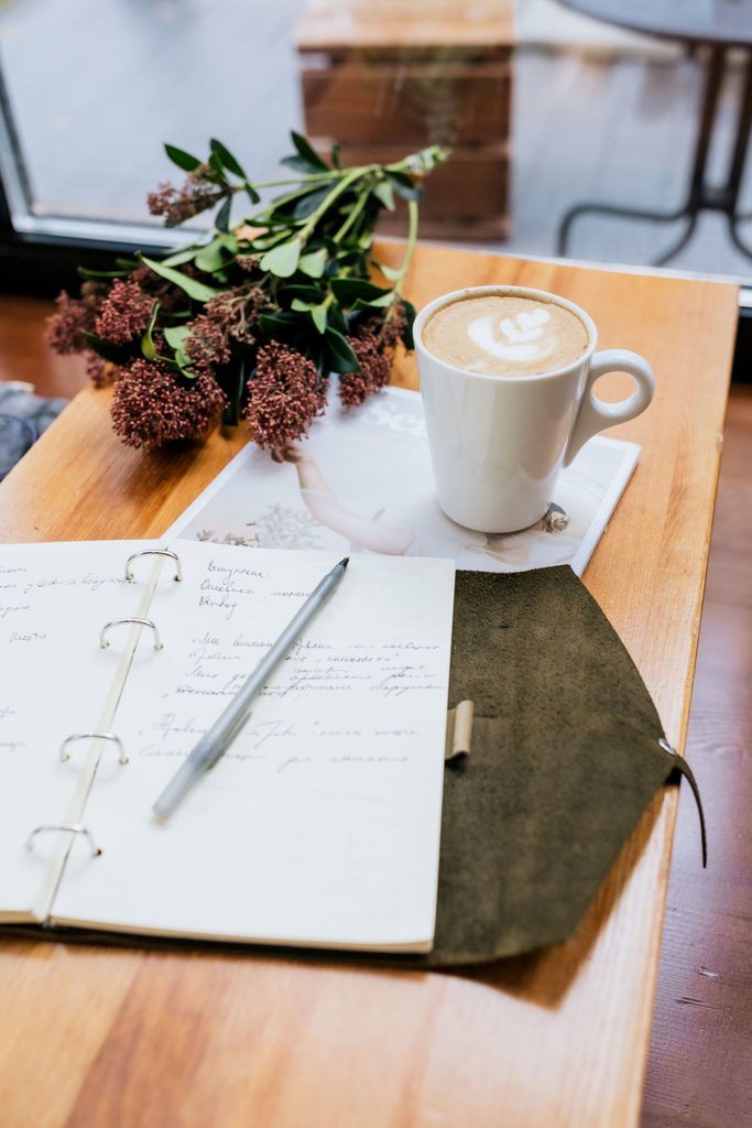Carnet de voyage manuscrit ouvert sur une table en bois, avec un café et des fleurs, ambiance d'écriture au calme