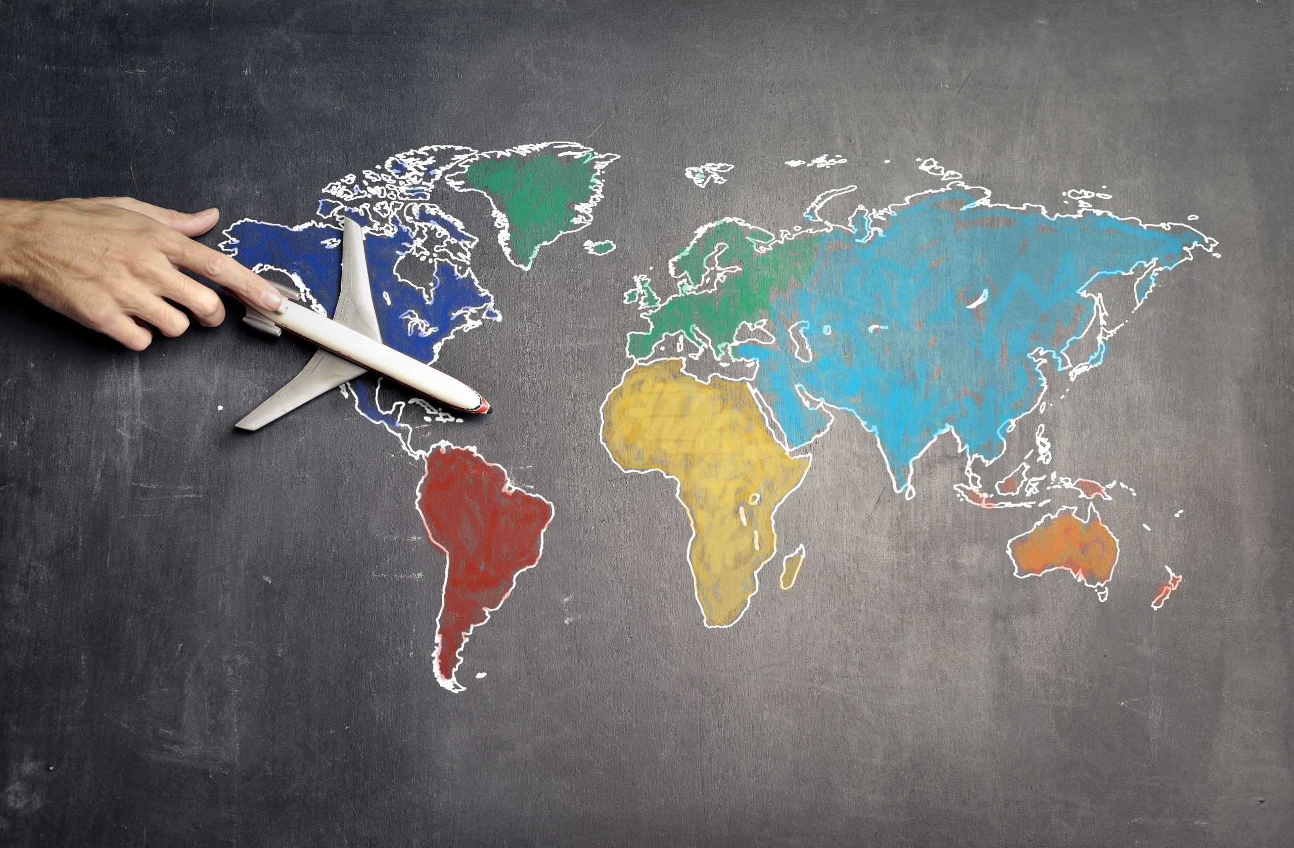 Main pointant une carte du monde dessinée à la craie avec les continents colorés et un avion miniature, 11 pays pas chers à découvrir