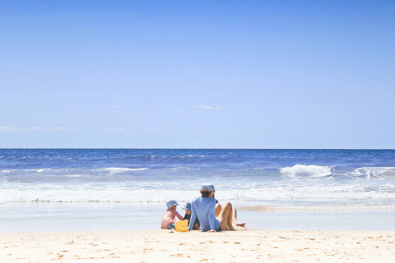 Parent et deux jeunes enfants jouant dans le sable avec un seau jaune au bord de la mer