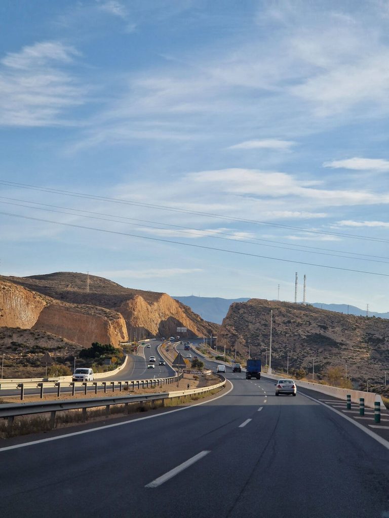 Autoroute ensoleillée traversant un paysage aride typique du sud de l'Europe, avec plusieurs voitures en circulation, pour un road trip pas cher