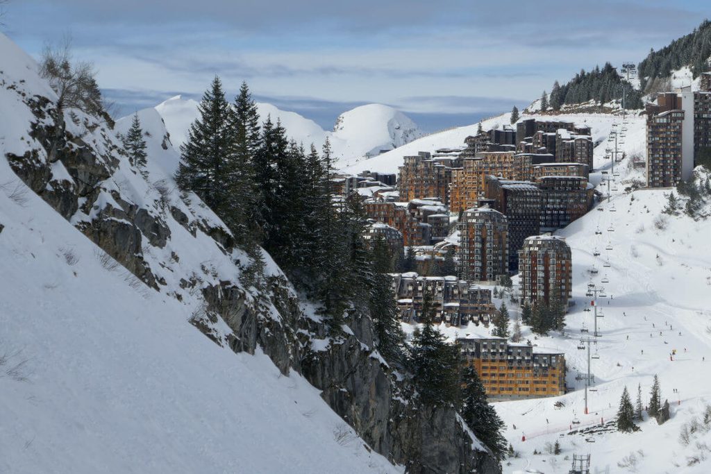 Village de montagne enneigé en France avec immeubles typiques et sapins, vue depuis les pistes de ski